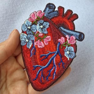 May include: An embroidered patch shaped like an anatomical heart in shades of red, featuring blue veins and arteries, decorated with light blue and pink flowers growing across the surface with detailed stitching and a dark border.