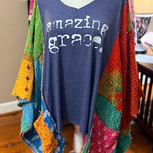 May include: A blue V-neck top with the words "amazing grace" in white. The sleeves and sides are patchwork with various colors and patterns, including red, green, yellow, and blue. The top is displayed on a mannequin.