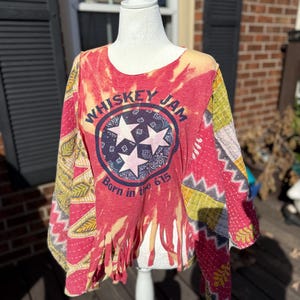 May include: A red, yellow, and blue tie-dye top with the text "Whiskey Jam" and "Born in the 615" printed on the front. The top has a round neck, long sleeves, and fringe detailing at the bottom. The sleeves have a patchwork design.