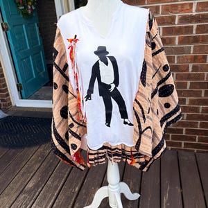 May include: A white V-neck shirt with a black silhouette of a man in a hat. The shirt is part of a poncho-style top with tan and black patterned fabric and orange fringe.