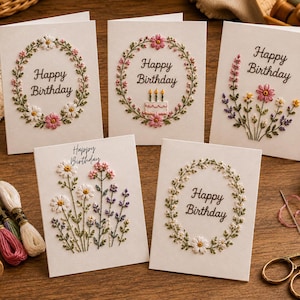 May include: Five white greeting cards featuring embroidered floral designs and the text Happy Birthday. The cards display wreaths of flowers, a small birthday cake, and wildflower bouquets in various colors including pink, white, and green.