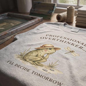 May include: A light grey heathered t-shirt featuring a graphic of a green frog wearing a straw hat, resting its chin on its hand. The text reads "PROFESSIONAL OVERTHINKER... I'LL DECIDE TOMORROW." A small dragonfly is illustrated next to the frog.