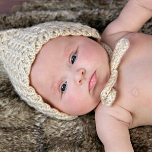Op de afbeelding: Een baby met een lichtbeige, gebreide muts met strikjes, liggend op een bruine en tan faux fur deken. De muts heeft een textuurpatroon en bedekt het hoofd en de oren van de baby.
