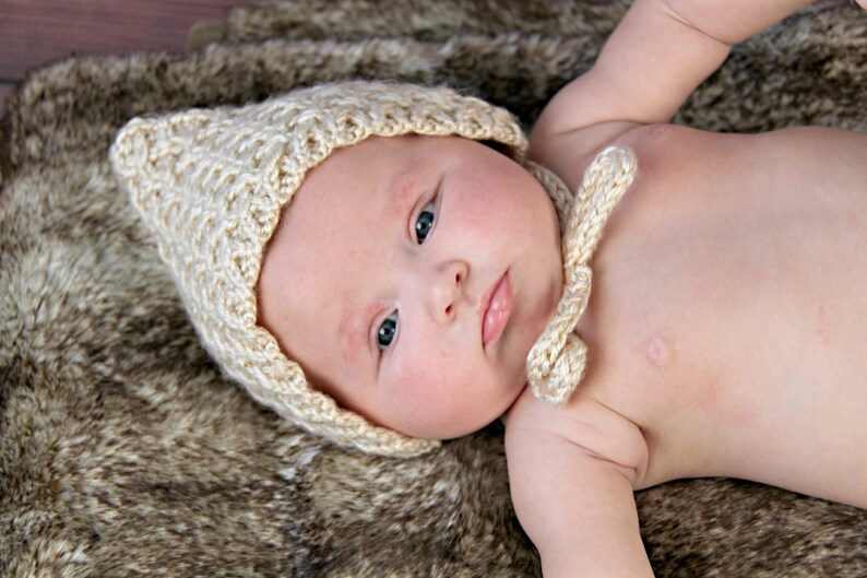 Op de afbeelding: Een baby met een lichtbeige, gebreide muts met strikjes, liggend op een bruine en tan faux fur deken. De muts heeft een textuurpatroon en bedekt het hoofd en de oren van de baby.