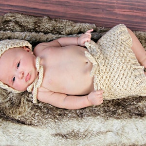 Op de afbeelding: Een pasgeboren baby draagt een cr&egrave;mekleurige gebreide outfit, inclusief een muts en een korte broek, en ligt op een imitatiebont deken. De baby ligt op een houten oppervlak, wat een zachte en natuurlijke uitstraling cre&euml;ert.