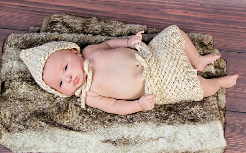 Op de afbeelding: Een pasgeboren baby draagt een cr&egrave;mekleurige gebreide outfit, inclusief een muts en een korte broek, en ligt op een imitatiebont deken. De baby ligt op een houten oppervlak, wat een zachte en natuurlijke uitstraling cre&euml;ert.
