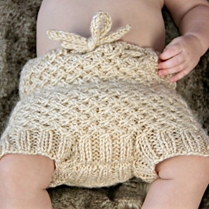 Op de afbeelding: Handgebreide babybloomers in een zachte, lichtbeige kleur. De bloomers hebben een textuursteekpatroon en een decoratieve strik in de taille. Het babykledingstuk is ontworpen voor comfort en stijl.