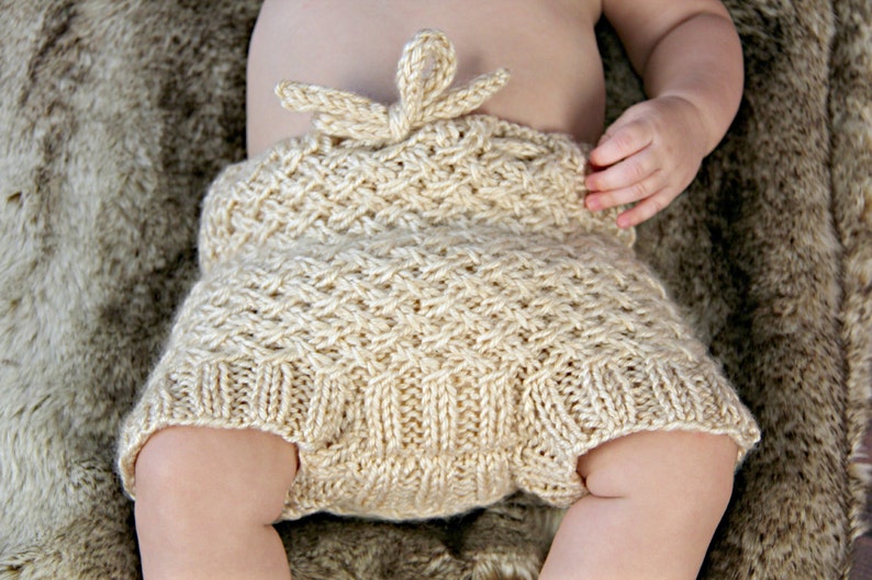 Op de afbeelding: Handgebreide babybloomers in een zachte, lichtbeige kleur. De bloomers hebben een textuursteekpatroon en een decoratieve strik in de taille. Het babykledingstuk is ontworpen voor comfort en stijl.