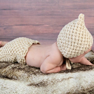Op de afbeelding: Een baby met een beige gebreide muts en luierbroekje, liggend op een imitatiebont kleed. De muts heeft een puntige bovenkant en het luierbroekje heeft een gerimpelde rand. De achtergrond is een houten oppervlak.