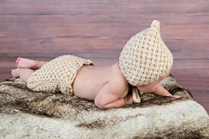 Op de afbeelding: Een baby met een beige gebreide muts en luierbroekje, liggend op een imitatiebont kleed. De muts heeft een puntige bovenkant en het luierbroekje heeft een gerimpelde rand. De achtergrond is een houten oppervlak.