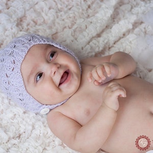 Peut inclure: Un bébé souriant portant un bonnet en dentelle violet clair avec un bouton, allongé sur une couverture blanche texturée. Le bébé porte un short blanc à volants. Portrait de studio.