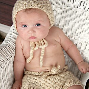 Peut inclure: Un bébé porte un ensemble beige tricoté, comprenant un chapeau pointu et un short, assis dans une chaise en osier blanche. L'ensemble présente un motif texturé et des détails de liens. Le bébé a la peau claire et les yeux bleus.