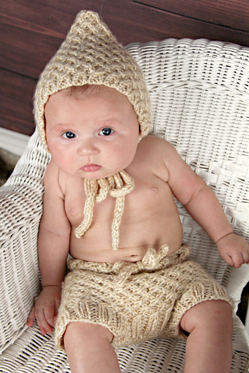 Op de afbeelding: Een baby draagt een gebreide beige outfit, bestaande uit een puntmuts en een korte broek, en zit in een witte rieten stoel. De outfit heeft een textuurpatroon en strikdetails. De baby heeft een lichte huid en blauwe ogen.