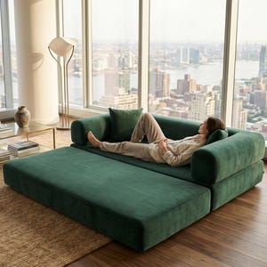 May include: A modern, emerald green sectional sofa with a chaise lounge. The sofa has a plush, rounded design and is positioned in front of a large window overlooking a cityscape. A matching pillow is on the sofa.