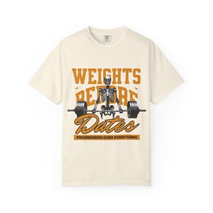May include: Cream-colored t-shirt featuring a skeleton lifting a barbell. The shirt has the words "WEIGHTS BEFORE DATES" in orange, with "PROGRESSION OVER EVERYTHING" below. A graphic tee for fitness enthusiasts.
