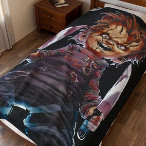 Può includere: Una coperta nera con una grafica di Chucky, la bambola assassina, che tiene due coltelli. La bambola ha i capelli rossi, cicatrici cucite e indossa una camicia a righe e una tuta. La parola "Good" è visibile sulla tuta.