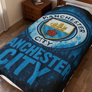 Puede incluir: Una manta azul y negra con el logotipo del Manchester City. El logotipo circular muestra un barco, una rosa roja y las palabras "Manchester City" en un estilo desgastado. La manta está sobre una cama.