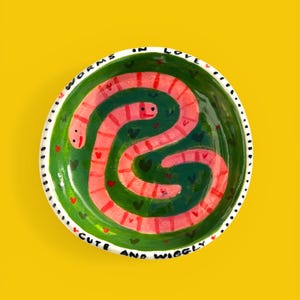 以下が含まれることがあります： 黒い持ち手と「But First, Coffee」のテキストが黒い太字で書かれた白いセラミックマグ。マグカップはぼやけた背景に置かれており、居心地の良い環境を示唆しています。