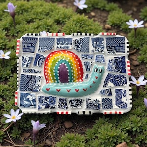 Peut inclure: Une sculpture de mosaïque en forme d'escargot fantaisiste, avec une coquille arc-en-ciel et un corps turquoise. L'escargot est décoré de cœurs rouges et repose sur une base de carreaux à motifs bleus et blancs. La bordure est blanche avec des accents noirs et rouges.