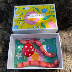 Op de afbeelding: Een witte rechthoekige doos met twee roze en rood gestreepte wormfiguurtjes met kleine gezichtjes en een rode paddenstoelhoed met witte stippen. Het deksel heeft een gele achtergrond met bloemen en de tekst Worms in love.
