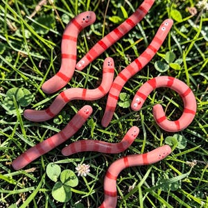 May include: A collection of seven small, smooth, pink and red striped glass worm figurines. Each worm features a simple, hand-painted face with two black dots for eyes and a small curved line for a mouth, arranged in various curved positions.