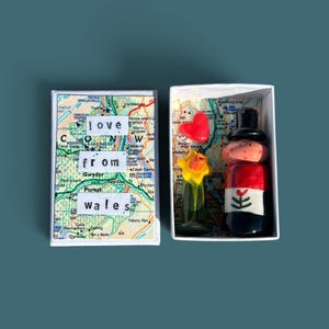 May include: A small white gift box containing a ceramic figure, a yellow daffodil, and a red heart. The box lid features a map of Wales with the text "love from wales" printed on white paper strips. The interior base is lined with a matching map.