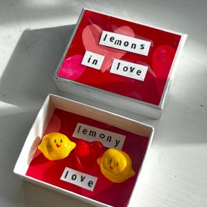 Könnte beinhalten: Zwei kleine, weiße Schachteln mit roten Innenräumen und Deckeln. Auf einer Schachtel steht "lemons in love" und auf der anderen "lemony love". Die zweite Schachtel enthält zwei gelbe, zitronenförmige Objekte mit lächelnden Gesichtern und einem roten Herzen.
