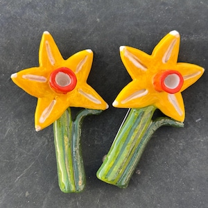 Peut inclure: Deux broches en forme de fleurs de jonquille. Chaque broche présente une fleur jaune vif en forme d'étoile avec un centre rouge et des accents blancs. Les tiges sont vertes avec des rayures jaunes.