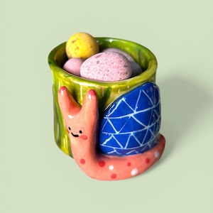 May include: A whimsical ceramic snail-shaped container. The snail has a peach-colored body with red and white polka dots, a blue shell with a white geometric pattern, and a green rim. The container holds several colorful, round candies.