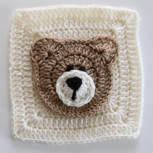 Può includere: Un quadrato all'uncinetto color crema con un motivo a testa di orso. La testa dell'orso è marrone con un muso bianco e un naso nero. Il quadrato ha un aspetto strutturato e fatto a mano, adatto per una coperta per bambini o decorazioni per la cameretta.