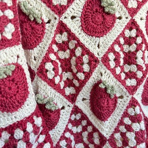 Puede incluir: Una manta de ganchillo hecha a mano con un patrón repetido de fresas rojas con hojas verdes sobre un fondo blanco. La manta está hecha de cuadrados unidos, creando un aspecto texturizado y acogedor.