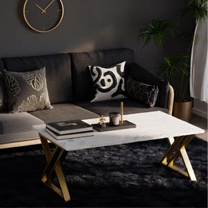 May include: A modern living room scene featuring a white marble-topped coffee table with gold-colored legs. The table is set on a black rug, with a dark gray sofa and decorative pillows. A gold-framed clock hangs on the wall.