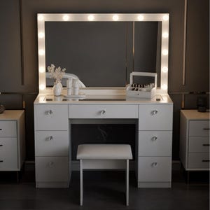 May include: A white vanity table with a large mirror framed by light bulbs. The vanity has multiple drawers, a stool, and a mirrored surface. A small white vase with flowers and a makeup organizer are on the vanity.