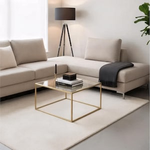 May include: A modern living room with a beige sectional sofa, a gold-framed glass coffee table, and a black tripod floor lamp. A dark gray throw blanket rests on the sofa. The room is decorated with a neutral color palette and a light-colored rug.