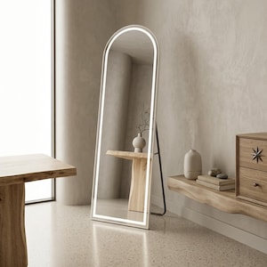 May include: A tall, arched floor mirror featuring a slim white frame with integrated LED lighting along the inner edge. The mirror stands on a dark metal support frame, reflecting a wooden console table with a vase and branches in a bright room.