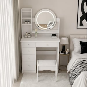 May include: A white vanity set with a round, illuminated mirror, a stool, and a built-in shelf. The vanity has drawers and a desk area. A white rug and a bed with a black and white blanket are in the background.