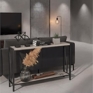 May include: A modern console table with a light wood top and black metal frame. The table is decorated with books, a small potted plant, and decorative objects. A clear glass vase with dried pampas grass sits below. A black floor lamp with a round bulb is in the background.