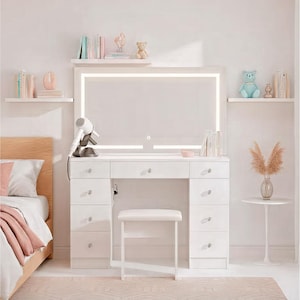 May include: A white vanity table with a large, illuminated mirror. The table has multiple drawers and a small stool. A hairdryer sits on the table. Shelves above hold books and decorative items. A bed is visible on the left.