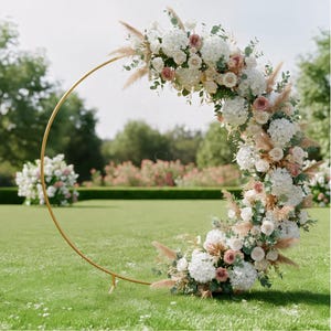 May include: A golden metal wedding arch adorned with white hydrangeas, blush roses, and pampas grass. The floral arrangement is arranged in a crescent shape, set on a green lawn with a blurred background of trees and flowers.