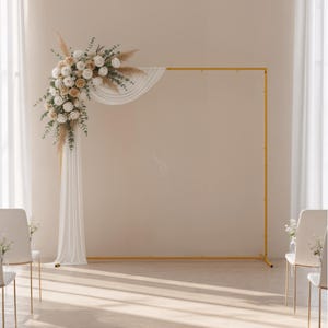 May include: A gold metal wedding arch decorated with white fabric and floral arrangements. The flowers are a mix of white and cream-colored roses, with greenery and pampas grass accents. Two white chairs with gold legs are in the foreground.