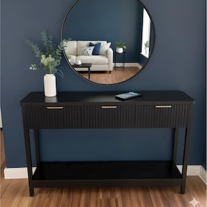 May include: A black console table with three drawers and gold-colored handles. The table has a ribbed texture on the drawer fronts and a lower shelf. A round mirror hangs above the table, reflecting a living room scene. A white vase with greenery sits on the table.