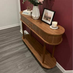 May include: A wooden console table with a curved design, featuring a drawer and a lower shelf. The table is adorned with a white vase, books, a framed picture, and a lit candle. The table is made of wood and has a warm, natural finish.