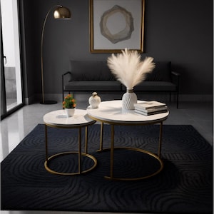 May include: Two white marble-topped coffee tables with gold metal frames. One table is smaller than the other. A black rug with a swirl pattern is beneath the tables. A vase with white fluffy plumes sits on the larger table.