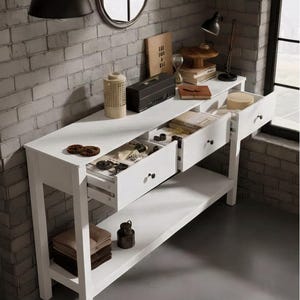 May include: A white console table with three drawers, open to reveal contents. The table has a lower shelf with folded textiles and a decorative jar. A brick wall and a black lamp are in the background.