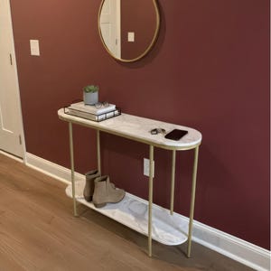 May include: A modern entryway table with a white marble top and a lower shelf. The table has gold-colored metal legs and a matching gold frame. A round mirror hangs on the burgundy wall above the table. The table is 48 inches long.