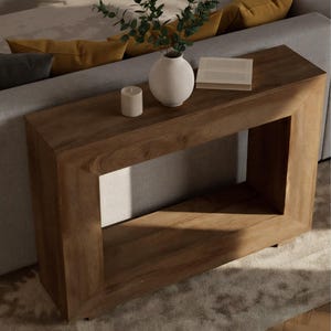 May include: A rectangular wooden console table with a natural finish. A white vase with greenery, a small white candle, and a white tray sit on top. The table is placed in front of a gray sofa with yellow pillows.