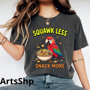 Peut inclure: T-shirt gris foncé avec un perroquet coloré. Le perroquet rouge, bleu et vert est au-dessus d'un bol de collations. Le texte dit "SQUAWK LESS SNACK MORE" en jaune et orange.