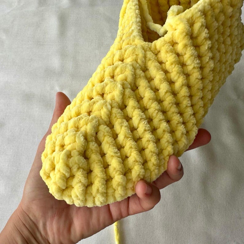 以下が含まれることがあります： 柔らかく質感のある糸で作られた、手作りの明るい黄色のスリッパ。 スリッパは手で持たれており、ふわふわで居心地の良い素材と詳細なかぎ針編みの作品を紹介しています。 スリッパは室内での使用を目的としています。