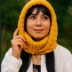 Könnte beinhalten: Nahaufnahme einer Person, die eine senfgelbe Strickhaube trägt. Die Haube ist aus dickem Garn gefertigt und hat eine gerippte Textur. Die Person trägt ein weißes Hemd mit Knöpfen und eine goldene Anhängerkette.