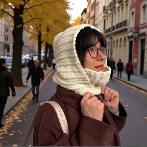 May include: A cream-colored, crocheted hood and cowl set. The hood frames the face, and the cowl wraps around the neck. The set is worn with a brown coat. The background shows a city street with people and autumn trees.